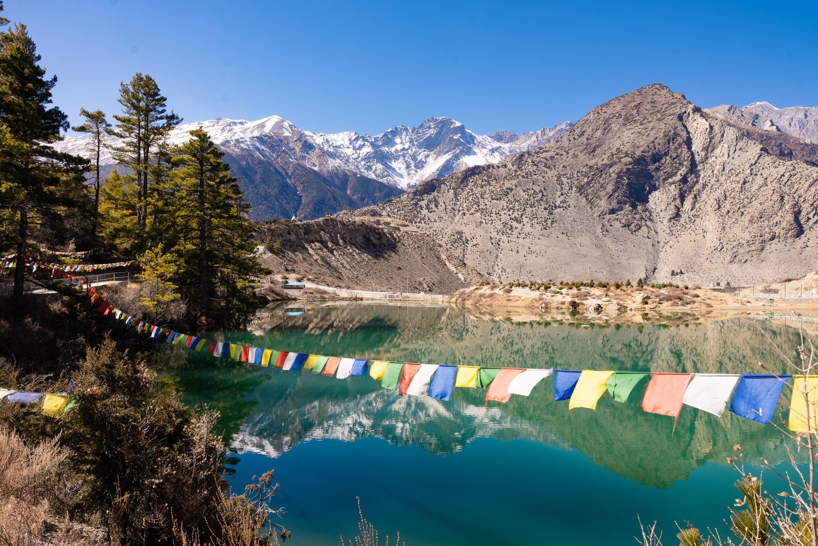 Scenic view of colorful prayer flags with Kagbeni's mountains and lake in Nepal, capturing natural beauty.